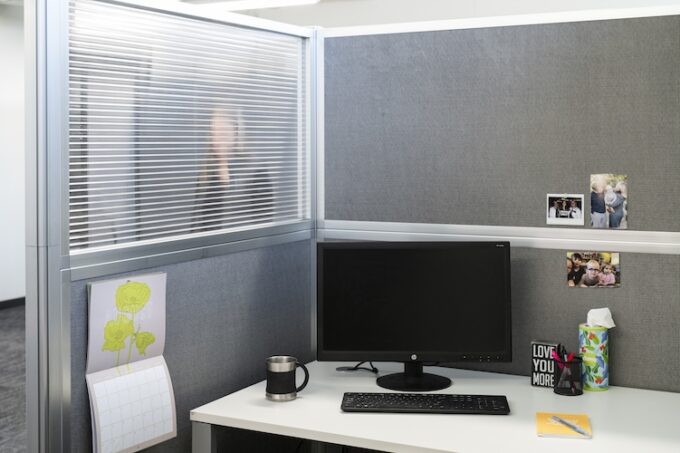 Office cubicle workstation with gray fabric panels and a fluted polycarbonate window, featuring a desk with a computer monitor, keyboard, mug, calendar, and personal photos.