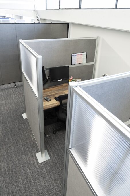 Modern office cubicle workspace with gray partition panels, a wooden desk, computer monitor, office chair, and small desk accessories