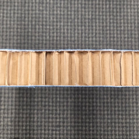 Close-up cross-section of an acoustic panel showing a layered core with folded cardboard structure and gray fabric outer surface.